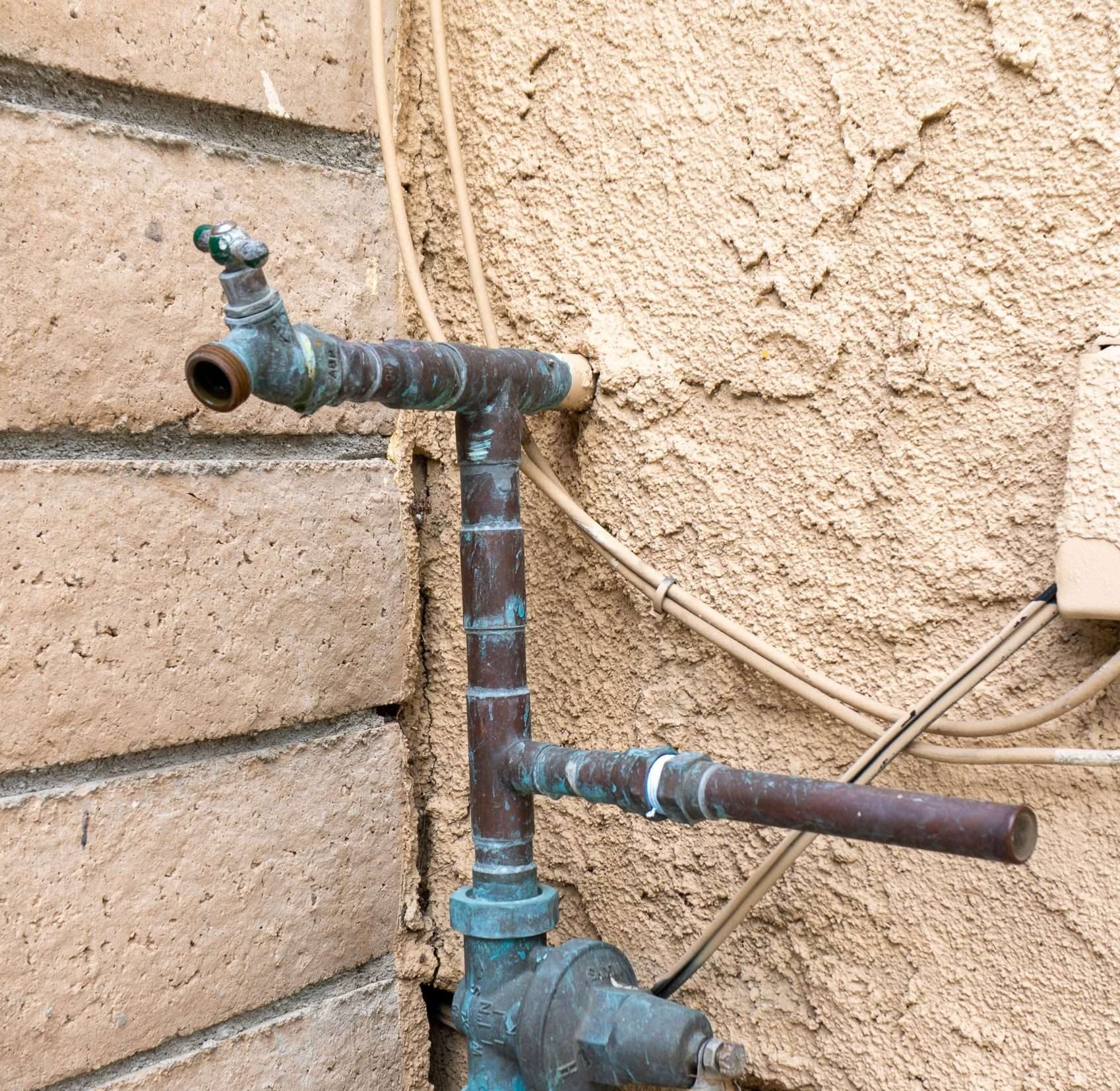 Aging copper outdoor hose bib and plumbing pipes showing corrosion and mineral buildup on exterior wall.